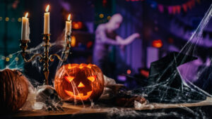 Halloween Still Life Colorful Theme: Scary Decorated Dark Room with Table Covered in Spider Webs, Burning Pumpkin, Candlestick, Witch's Hat and Skeleton. In the Background Silhouette of Monster Walking By